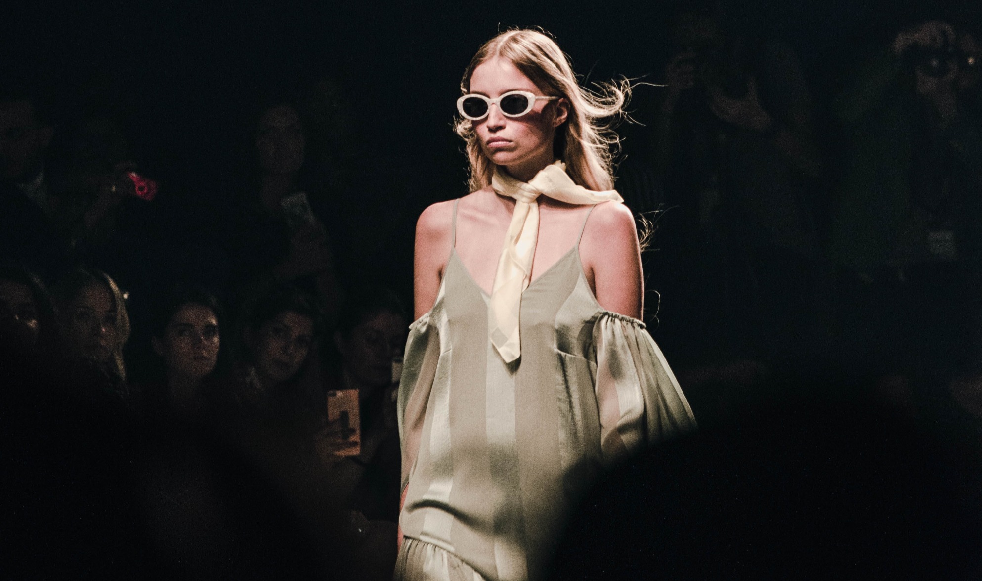 A grumpy looking fahsion model in large sunglasses and a beige dress walks down a catwalk